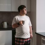 Young man with Down syndrome holding a cup, enjoying a quiet moment by a window in a modern kitchen.