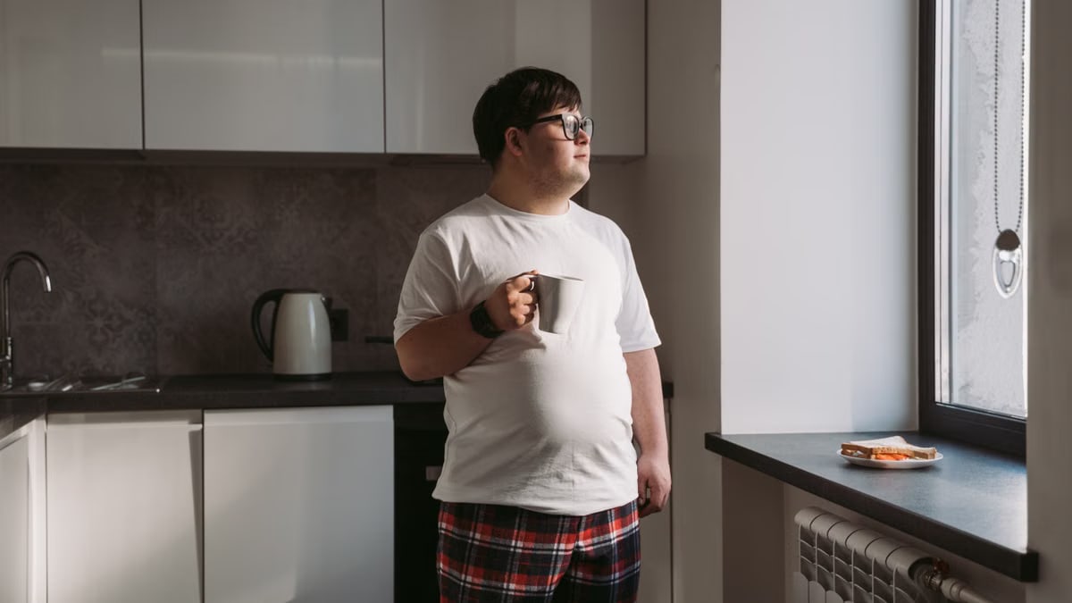 Young man with Down syndrome holding a cup, enjoying a quiet moment by a window in a modern kitchen.